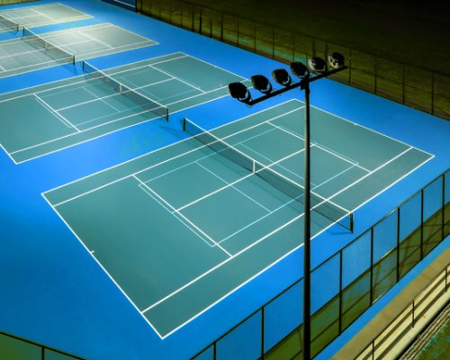 Evening aerial photo of outdoor blue tennis courts with pickleball lines with lights turned on.