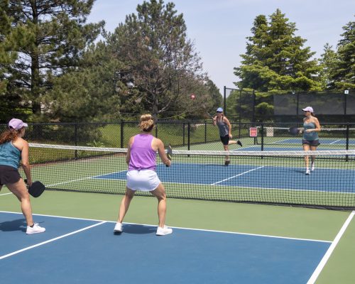 A pickleball player serves the ball during a game of doubles on a suburban court during summer.